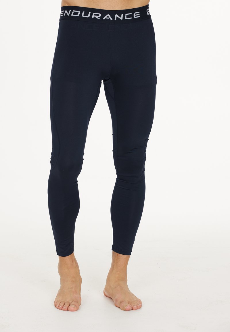 Mand iført marineblå kompressionsleggings med "ENDURANCE" på linningen, stående barfodet mod hvid baggrund.