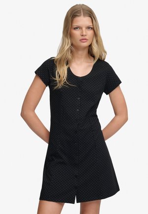 Młoda kobieta o długich blond włosach, ubrana w czarną sukienkę na krótki rękaw z małymi białymi kropeczkami, stoi z rękami za plecami.