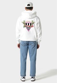 Biała bluza z kapturem z kolorowym wzorem na plecach, zawierającym wzory, liście palmowe i trójkątny kształt. W połączeniu ze jasnoniebieskimi dżinsami.