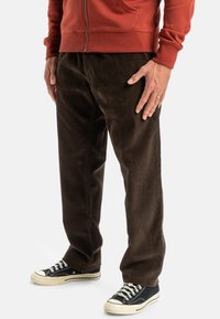 Brown corduroy pants with a relaxed fit, paired with black sneakers featuring white accents. The fabric shows vertical ribbing texture.