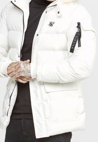 Vit pufferjacka med huva, framdragkedja och elastiska ärmslut. Har en ficka med dragkedja på ärmen och en stor framficka.