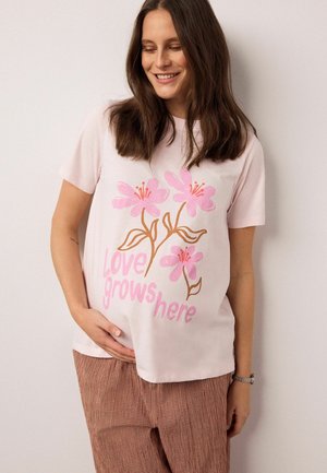 Lyserød bomulds-t-shirt med tre lyserøde blomster og teksten "Love grows here" i fede lyserøde bogstaver, kombineret med brune ternede bukser.