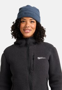 Lachend persoon met krullend haar, draagt een blauwe Jack Wolfskin fleece muts en een zwarte structuur gehalveerde fleece jack.