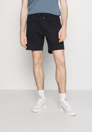 Mann trägt schwarze Shorts mit Kordelzug, weiße Socken und weiße Turnschuhe, steht vor einem schlichten hellen Hintergrund.