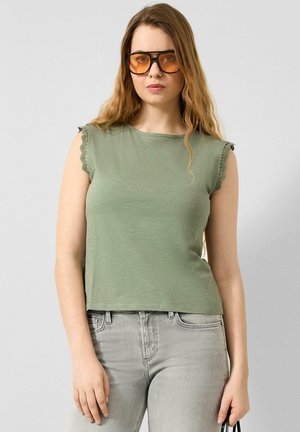 Vrouw met lang golvend haar, die oranje getinte zonnebril draagt, een olijfgroene mouwloze top met kanten rand, en lichtgrijze spijkerbroek, terwijl ze een zwarte tas vasthoudt.