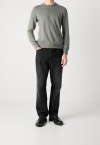 Mies, joka on pukeutunut oliivinvihreään pitkähihaiseen paitaan, mustiin farkkuihin ja mustiin nahkakenkiin, seisoo yksi käsi taskussa valkoista taustaa vasten.