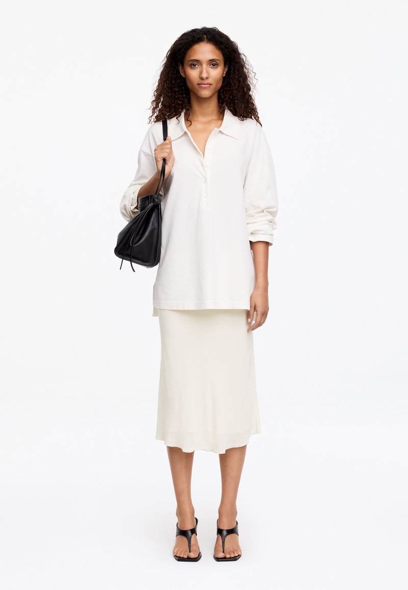 Femme aux cheveux bouclés porte une chemise blanche oversize, une jupe midi crème, des sandales à talons noires, et tient un sac à main noir à l'épaule.