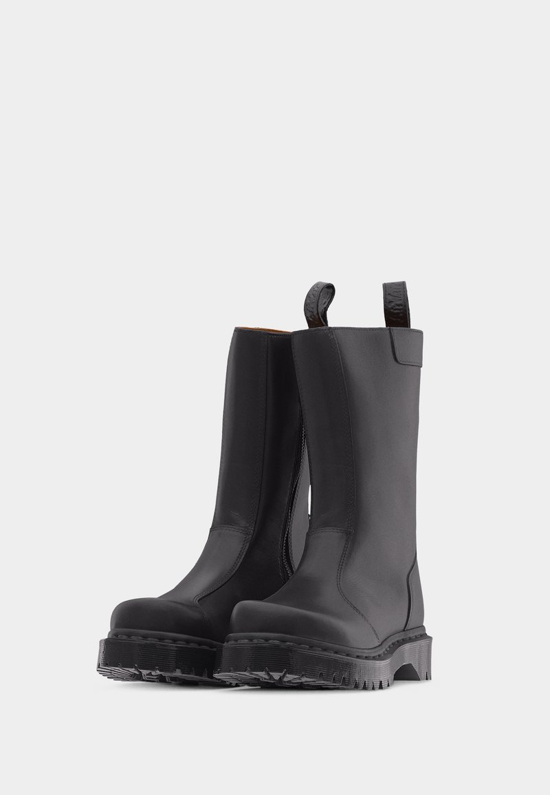 Coppia di stivali neri in pelle fino a metà polpaccio con suole spesse a banda larga e linguette per infilarsi, in piedi su uno sfondo grigio chiaro uniforme.