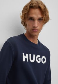 Felpa navy in cotone, caratterizzata da un collo rotondo e un audace logo bianco "HUGO" stampato sul davanti. Tessuto morbido e vestibilità rilassata.
