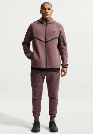 Mężczyzna stojący, ubrany w fioletowo-różowy dres Nike z kurtką na zamek i spodnie dresowe, czarne trampki, ręce rozluźnione, neutralne tło.