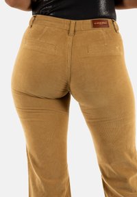Pantalones de pana de tiro alto en color mostaza con textura acanalada, que cuentan con bolsillos traseros y una etiqueta de cuero marrón con la marca en la cintura.