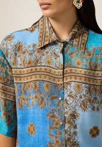 Niebieska koszula w wzory z motywami paisley i kwiatowymi w odcieniach brązu i złota. Klasyczny kołnierzyk zapinany na guziki. Gładka faktura tkaniny.