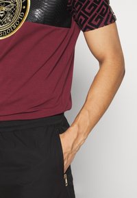 Bordeaux t-shirt met gestructureerde zwarte slangenpatroonaccenten op de schouders en een gouden embleem. Zwarte elastische shorts met ritszakdetail.