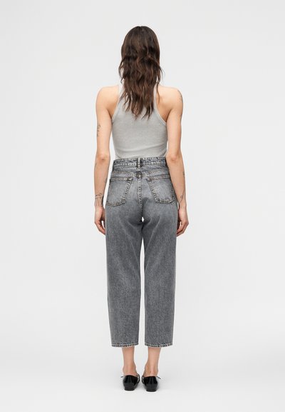 Γυναίκα με μακριά καστανά μαλλιά που φοράει γκρι αμάνικη μπλούζα, ψηλόμεσα γκρι τζιν και μαύρα παπούτσια, στέκεται με την πλάτη στο λευκό φόντο.