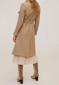 Manteau en laine beige avec taille ceinturée, coupe longueur genou, fente au dos, associé à une jupe plissée rose clair et des bottes à imprimé serpent.