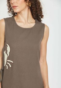 Mujer con cabello rizado que lleva un vestido sin mangas color topo con diseño blanco de cangrejo y pendiente texturizado dorado en forma de hoja, sobre fondo liso.