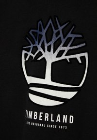 T-shirt nera in tessuto con logo grande bianco e blu sfumato di Timberland e la scritta "Timberland the original since 1973."