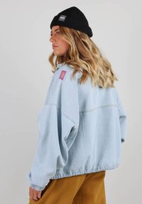 Veste en denim bleu clair avec une coupe oversize, des poignets élastiques et un patch logo violet sur l'épaule. Tissu texturé et design décontracté.