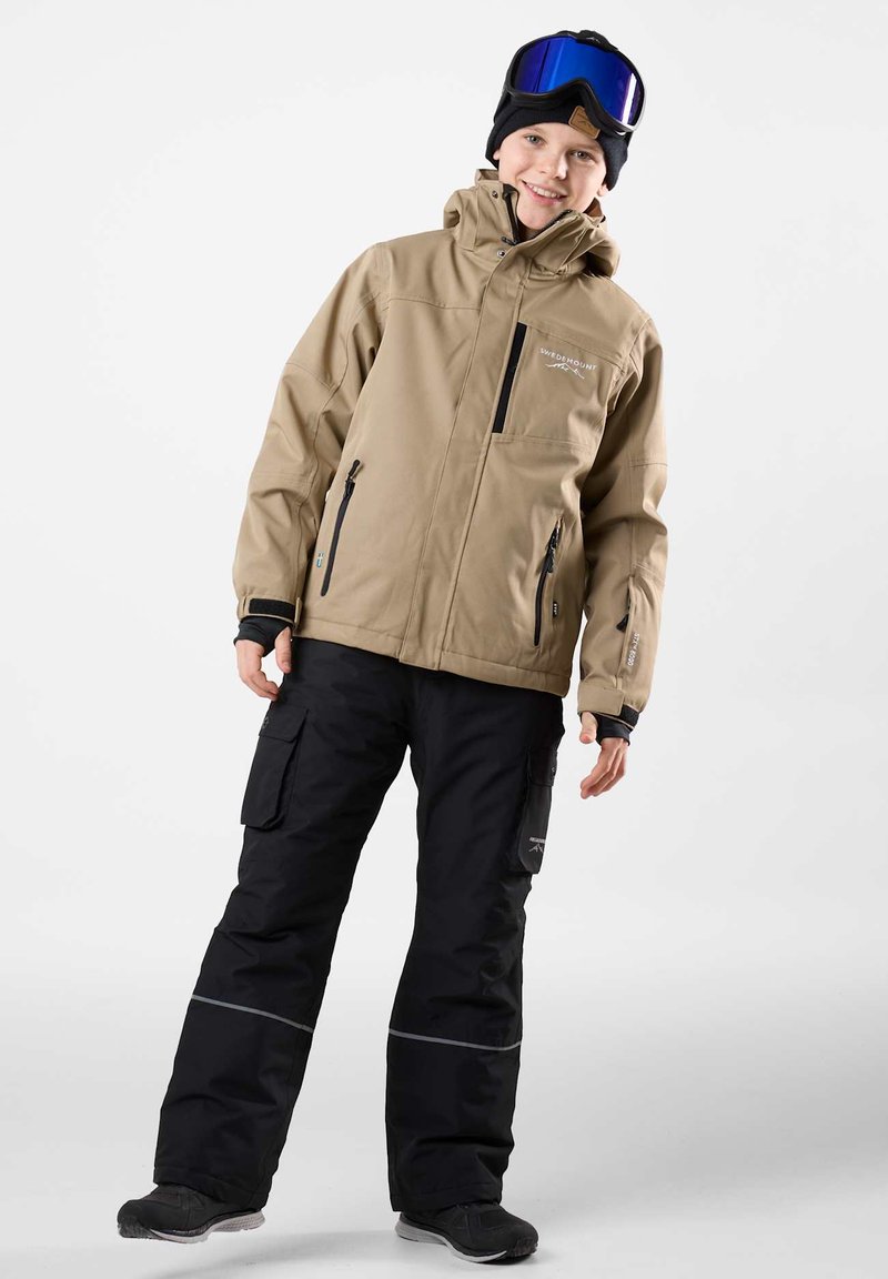 Beige wasserdichte Jacke mit einem Frontreißverschluss und Taschen, kombiniert mit schwarzen Schneehosen, die über Cargo-Taschen und reflektierende Streifen verfügen.