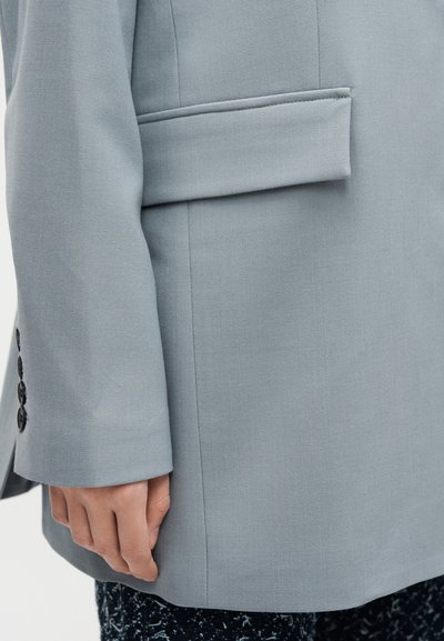 Μανίκι γαλάζιου σακακιού και πλαϊνή τσέπη με τρία μαύρα κουμπιά εμφανή στη μανσέτα, φορεμένα πάνω από μαύρο παντελόνι με σχέδια.