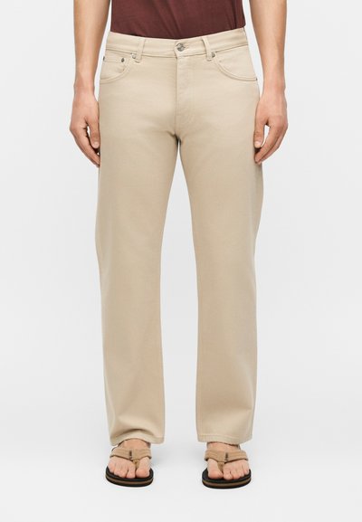 Homme portant un pantalon beige droit avec poches avant et sandales marron, debout devant un fond blanc.