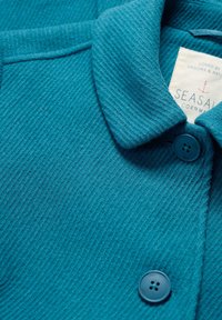 Veste texturée teal avec col et deux boutons bleus, présentant une étiquette blanche visible avec un logo d'ancre rouge et du texte à l'intérieur de l'encolure.