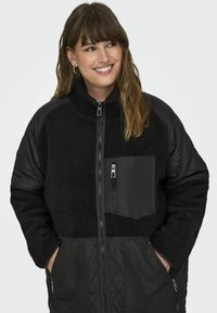 Black jacket with quilted sleeves and a fleece body. Features a zip front, high collar, and an exterior chest pocket with a zipper.