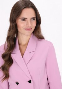 Blazer rosa chiaro con design doppiopetto, rever affilati e bottoni neri a forma di cuore, tessuto morbido con vestibilità strutturata.