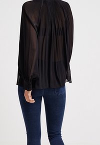 Blusa nera e trasparente con dettagli plissettati sul retro, maniche lunghe e polsini con bottoni, abbinata a jeans skinny blu scuro.