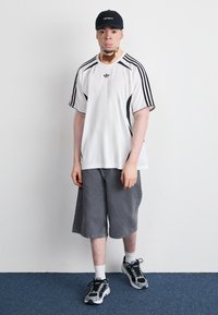 White athletic shirt with black accents and three stripes, gray shorts, and black sneakers with reflective details; worn with a black cap.