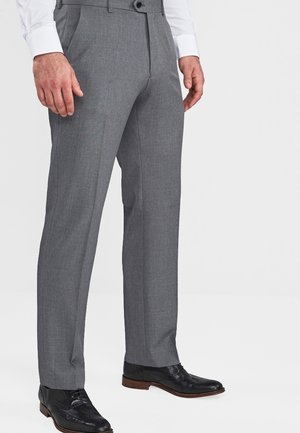 Mann trägt graue formelle Hose, schwarze elegante Schuhe und ein weißes langärmeliges Hemd, steht vor schlichtem weißem Hintergrund.