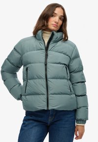 Polstret jakke i dæmpet teal, med lynlås fortil, høj krave og sidelommer. Glat tekstur med synlige sømme og en struktureret form.