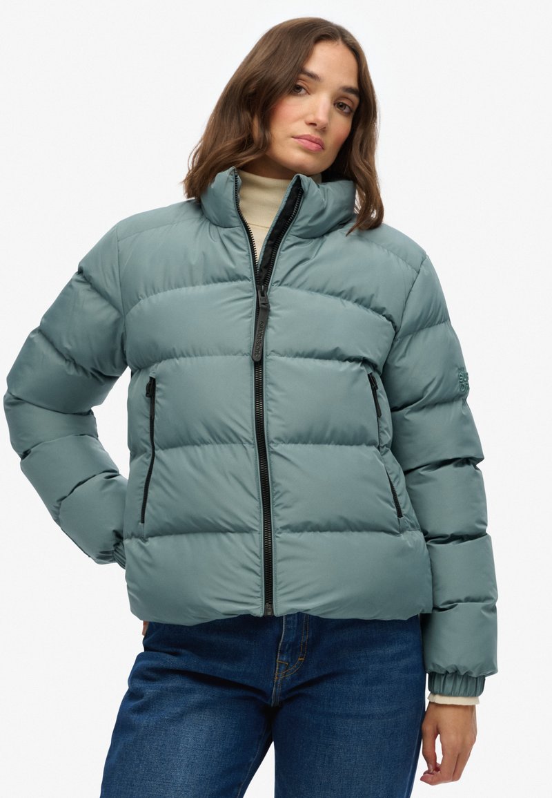 Polstret jakke i dæmpet teal, med lynlås fortil, høj krave og sidelommer. Glat tekstur med synlige sømme og en struktureret form.