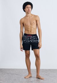 Mustad ujumispüksid punase nööriga, millel on valge "DIESEL" logo. Modell on palja ülakehaga, seisab tekstuuriga pinnal.