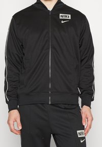 Giacca nera con zip realizzata in materiale liscio, con strisce bianche ai lati e un piccolo logo Nike sul petto e sulla tasca sinistra.