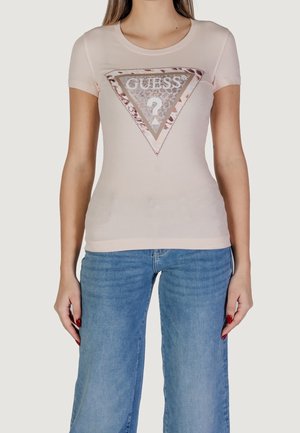 Lichtroze T-shirt met een metalen driehoeksgrafiek, met de tekst "GUESS" en een vraagteken; korte mouwen en een aangesloten pasvorm. Gecombineerd met een blauwe spijkerbroek.