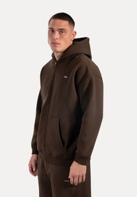 Brauner Reißverschluss-Hoodie mit Fronttasche, gerippten Bündchen und relaxed Fit. Mit kleinem Logo auf der Brust. Texturierte Stoffoberfläche.