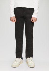 Pantaloni neri slim-fit in cotone, dotati di tasche anteriori, abbinati a sneakers bianche. L'outfit è completato da una camicia bianca a maniche lunghe.