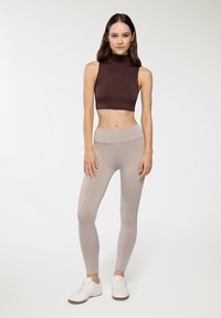 Ciemnobrązowy, bezrękawnikowy top typu crop paired z jasnobeżowymi legginsami o wysokim stanie. Model nosi białe sneakersy z teksturowaną podeszwą.