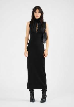 Black sleeveless dress with a high neck, featuring a keyhole cutout with buttons. Paired with knee-high black heeled boots. Looking straight ahead.