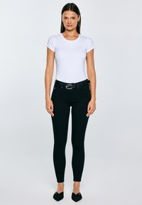 Sieviete stāv ar seju pret priekšu, valkājot baltu pieguļošu īsroku krekliņu, melnas augstajiem vidukļiem plānas džinsa bikses ar jostu un melnas smailas kurpes.