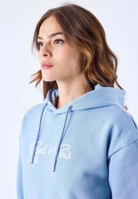 Felpa con cappuccio di colore azzurro chiaro, realizzata in tessuto morbido. Presenta un logo ricamato sul petto e terminali delle stringhe in tono argento.