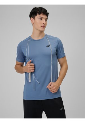 Joven con una camiseta azul 4F y pantalones negros, sosteniendo una cuerda para saltar colgada alrededor de su cuello, mirando hacia un lado.