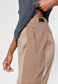 Καφέ αθλητικό σορτς με υφή, πλαϊνή τσέπη με φερμουάρ και κεντημένο λογότυπο "TERREX" στο κάτω μέρος. Φορεμένο με γκρι μπλούζα.