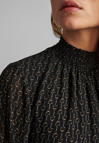 Blusa nera con colletto alto arricciato. Presenta un motivo ripetuto di forme geometriche dorate e bianche su un tessuto trasparente.
