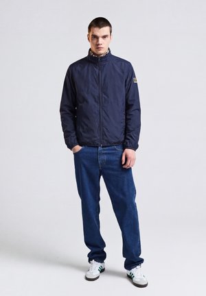 Junger Mann steht, trägt eine marineblaue Jacke mit Reißverschluss, blaue Jeans und weiße Sneaker mit grünen Streifen vor einfarbigem Hintergrund.