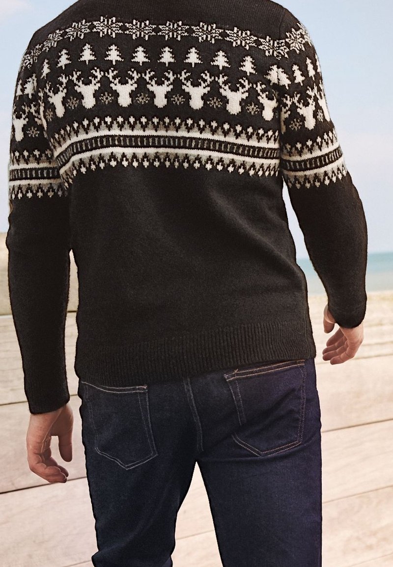 Suéter de punto negro con patrones geométricos blancos, que incluyen renos y copos de nieve, con puños y dobladillo acanalados, combinado con jeans azules.