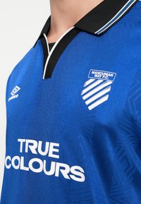 Mėlyna futbolo marškinė su juodu ir baltu apykakle, Mancunian Way F.C. logotipas, Umbro logotipas, ir rėmėjo tekstas "TIKRIEJI SPALVOS".