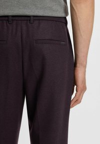 Mörk lila byxor i mjukt tyg, med en slank design, två bakfickor och en liten etikett på midjebandet.