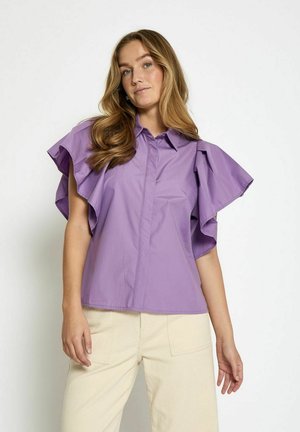 Purple short-sleeve blouse with large ruffle sleeves, collared design, and button front. Worn with light-colored, wide-leg trousers.